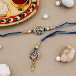 Royal-themed Bhaiya-Bhabhi Rakhi set with blue thread, crown and teardrop motif, and decorative kumkum-chawal thali with festive pearls and stones.