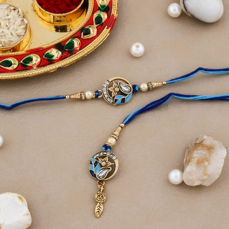 Royal-themed Bhaiya-Bhabhi Rakhi set with blue thread, crown and teardrop motif, and decorative kumkum-chawal thali with festive pearls and stones.